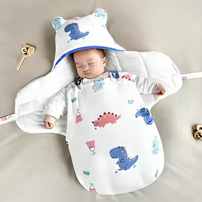 Allongé calmement, un bébé dort dans une gigoteuse hiver bebe blanche à imprimés de petits dinosaures, avec une capuche aux oreilles trop mignonnes.