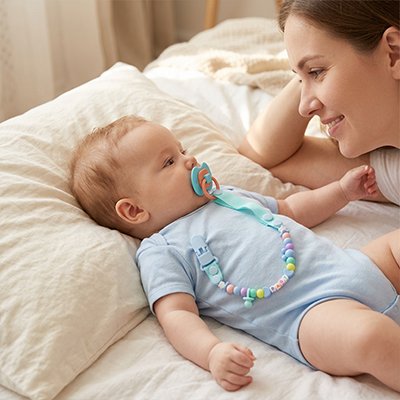Allongé sur le dos, bébé observe sa maman à ses côtés, sa tétine en bouche et fixée à son vêtement par un attache tétine SnapLoop™ bleue