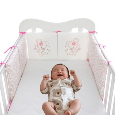 Bébé allongé sur son dos dans son lit entoure d'un doux tour de lit bébé fille blanc aux motifs fleuris et agrémenté de deux coussins brodés représentant un ourson