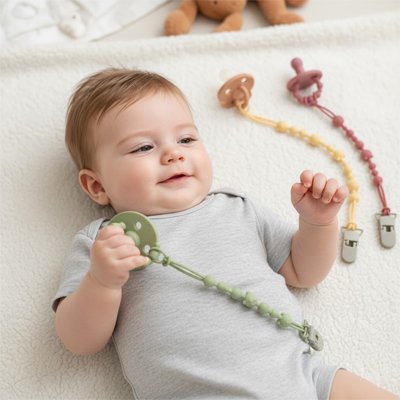 Bébé allongé sur le lit avec des attache tetines en silicone, verte, rose et jaune OoneyClip™