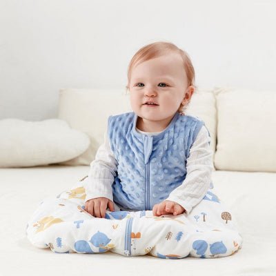 Bébé assis, souriant sur le lit, vêtu d’une turbulette hiver, bleue matelassée en haut et blanche avec des motifs d’animaux en bas