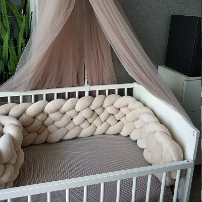Dans un lit bébé blanc, un tour de lit tressé beige apporte douceur et confort sublimé par un ciel de lit rose poudre.