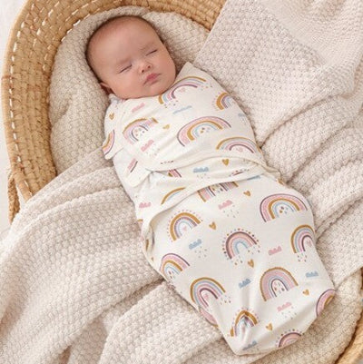Paisiblement endormi, le bébé repose dans un couffin en osier, enveloppé dans une turbulette blanche aux motifs arc-en-ciel délicats