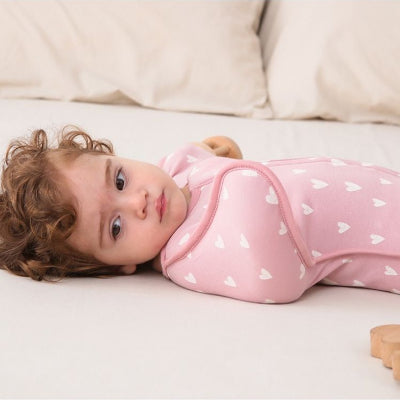 Sur un lit blanc, un bébé repose calmement, enveloppé dans sa turbulette gigoteuse rose aux jolis cœurs