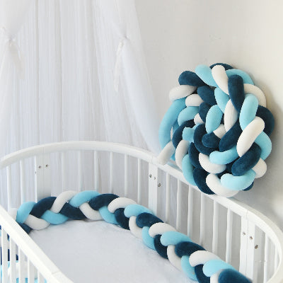 Un tour de lit tresse aux tons bleu, blanc et turquoise est installé dans un lit bébé blanc avec une seconde tresse enroulée et posée contre le mur sous un voile léger.