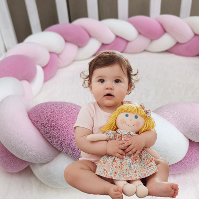 Un bébé assis dans son lit tenant tendrement une poupée est entouré d'une tresse tour de lit aux douces teintes rose et blanc