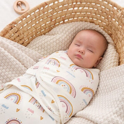 Un bébé dort paisiblement dans un couffin en osier, emmailloté dans une turbulette blanche ornée de motifs arc-en-ciel