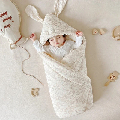 Un bébé dort paisiblement sur un lit blanc, enveloppé dans une turbulette à motif de forêt fantastique.