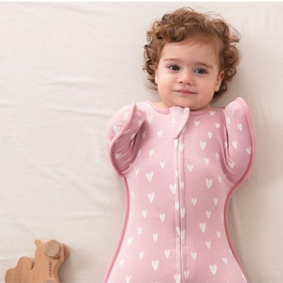 Un bébé dort paisiblement sur un lit immaculé, emmitouflé dans une turbulette gigoteuse rose ornée de motifs en forme de cœurs