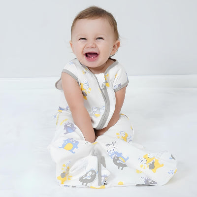 Un bébé est assis souriant, enveloppé dans une gigoteuse turbulette blanche à motifs de monstres amusants