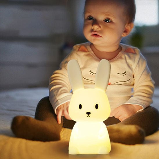 Un bébé est assis sur le tapis, avec devant lui sa veilleuse bébé Glowbit™ en forme de lapin lumineux.