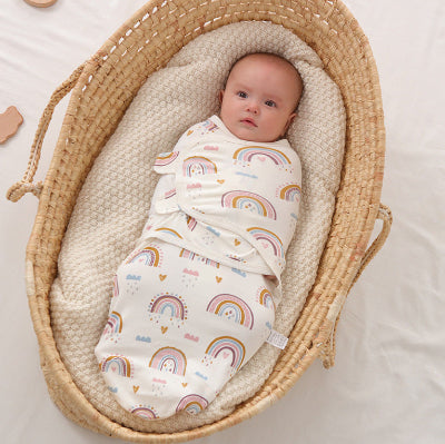 Un bébé se repose calmement dans un panier en osier, enveloppé dans une turbulette claire décorée de délicats motifs arc-en-ciel.