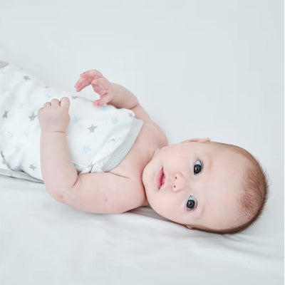 Un bébé repose paisiblement sur un lit blanc, emmailloté dans une gigoteuse emmaillotage aux motifs d’étoiles.