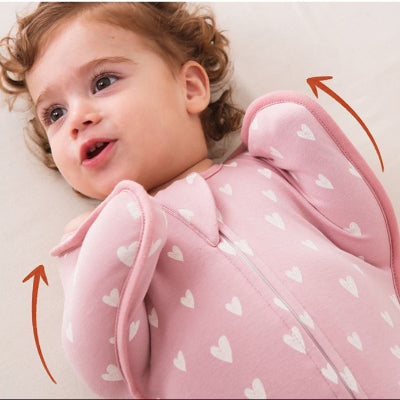 Un bébé repose sereinement sur un lit blanc, emmitouflé dans une turbulette gigoteuse rose parsemée de cœurs.
