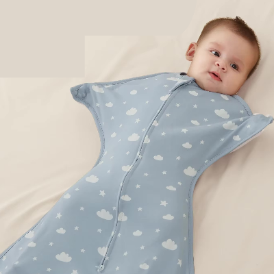 Un bébé repose tranquillement sur un lit, enveloppé dans une turbulette gigoteuse bleue décorée de nuages et d’étoiles
