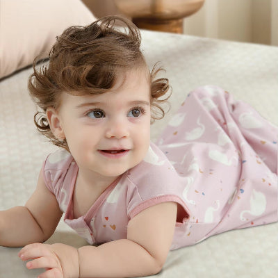 Un bébé souriant est allongé confortablement sur un lit, vêtu d’une gigoteuse bébé rose sans manches ornée de motifs cygnes.