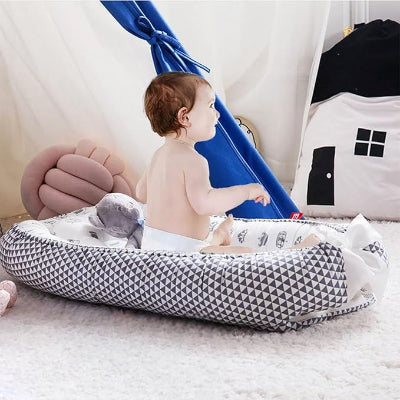 Un bébé souriant, assis dans son Berceau Bébé NidPortableBébé™ Couronne  déposé sur le tapis, avec son doudou à ses côtés.