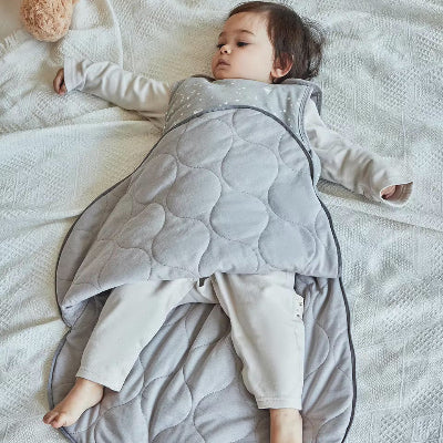 Un enfant est allongé sur le lit, bien au chaud dans une gigoteuse grise à motifs d’étoiles et de montgolfières