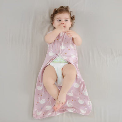 Une fillette est allongée sur un lit, portant une gigoteuse bébé rose sans manches ornée de motifs cygnes