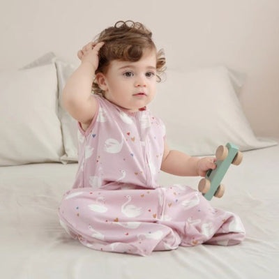 Une fillette est assise dans son lit, tenant une petite voiture en bois verte dans sa main, vêtue d’une gigoteuse bébé rose sans manches aux motifs de cygnes.