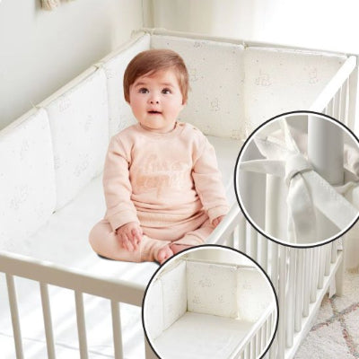 Une fillette est assise dans son lit, entourée d’un tour de lit bébé complet blanc rembourré orné de motifs d’éléphants, de petites étoiles et de lignes à petits points, qui enveloppe le lit de douceur et de sécurité.