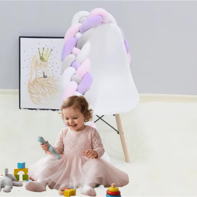 Une fillette joue avec ses jouets assise devant une chaise blanche sur laquelle est déposé un tour de lit tresse aux teintes pastel rose, blanc, gris et lilas