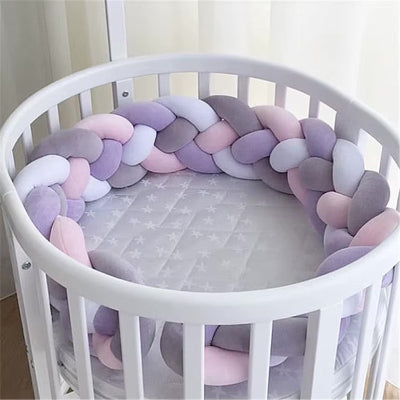 Un lit bébé rond blanc est délicatement entouré d'un tour de lit tresse aux teintes pastel rose, gris blanc et lilas, créant une atmosphère douce et apaisante