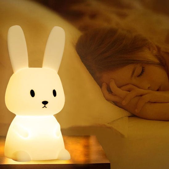 Une petite fille assoupie dans son lit douillet, veillée par la tendre lueur d'une veilleuse bébé Glowbit™ lapin posée près d'elle