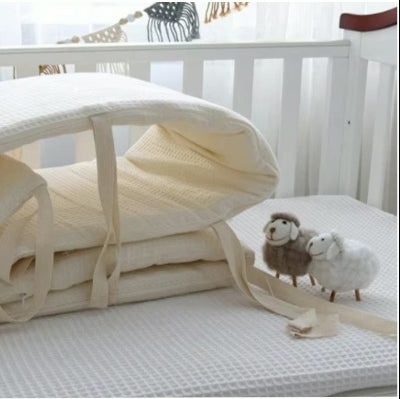 Un tour de lit bébé complet moelleux et en tissu gaufré beige est disposé dans le lit bébé accompagne de moutons en bois.