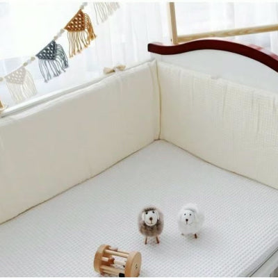 Un tour de lit bébé complet en tissu gaufre beige entoure le lit où sont poses de petits jouets en bois dont deux moutons.