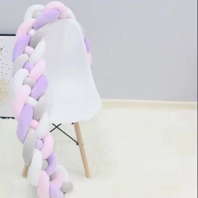 Un tour de lit tresse aux teintes pastel rose, blanc, gris et lilas est délicatement posé sur une chaise blanche, apportant une touche décorative douce et élégante.