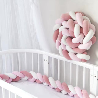 Une tresse tour de lit aux douces teintes rose et blanc entoure le lit bébé blanc tandis qu'une seconde tresse enroulée en boule est fixée au mur