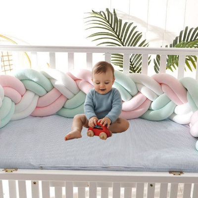 Une tresse tour de lit rose pastel blanche et vert menthe installée dans un lit bébé blanc tandis qu'un enfant assis joue avec sa voiture rouge diffuse une ambiance douce et apaisante.